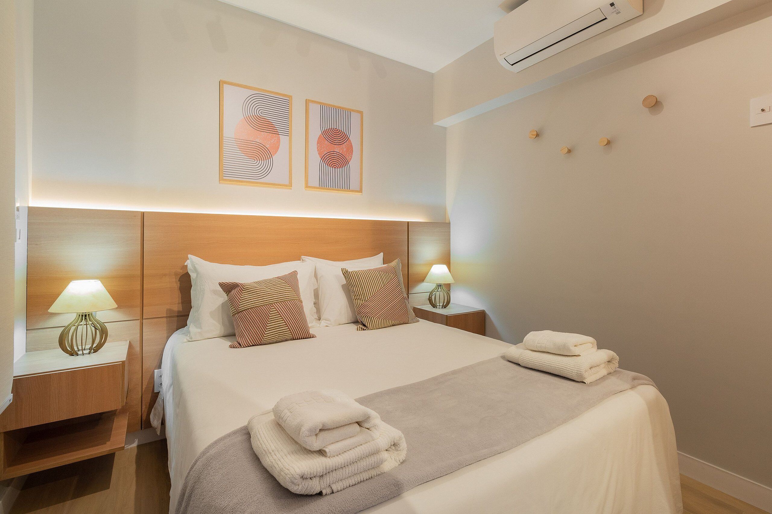 A neatly made bed with a folded towel, a bedside lamp, and a wall-mounted air conditioner.