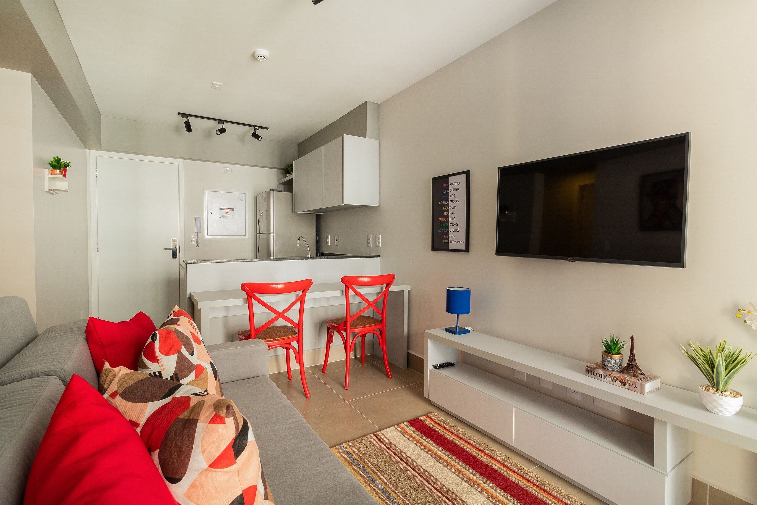 A modern living room with a grey sofa, red chairs, a flat-screen TV, and a kitchen in the background.