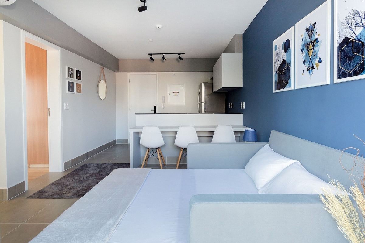 Moderni hotellihuone, jossa on sänky, oleskelualue, pieni keittiönurkkaus ja taideteoksia seinillä.