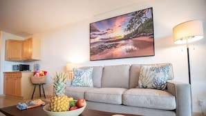 A living room with a grey sofa, a wooden coffee table, a bowl of fruit, and a painting of a beach scene above the sofa. - Perfect location in Kihei! 5 minutes to the beach! (Kihei)