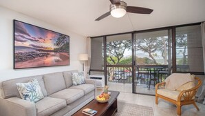 A living room with a sofa, coffee table, and armchair, a ceiling fan, a large window with a view of the outdoors, and a painting on the wall. - Perfect location in Kihei! 5 minutes to the beach! (Kihei)