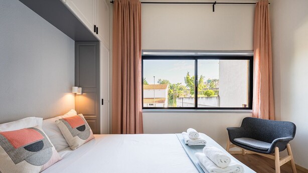 Hotellihuone, jossa on sänky, tuoli, ikkuna verhoilla ja näkymä ulkona oleviin rakennuksiin.