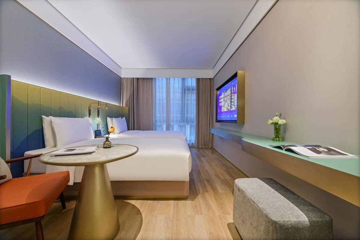 A hotel room with a bed, a desk, a chair, a small table, a TV, and a vase with flowers.