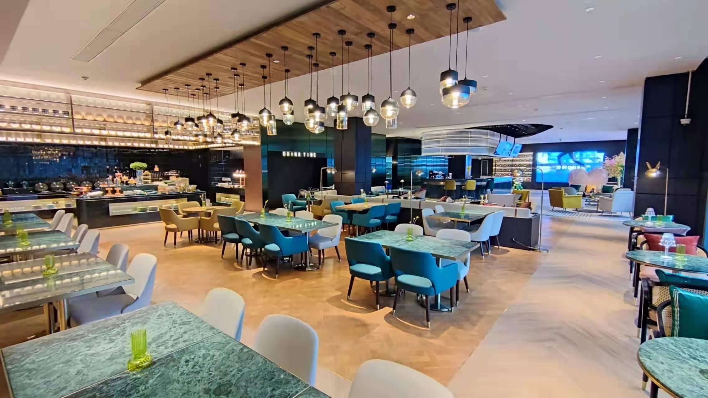 A modern restaurant with multiple dining areas, hanging pendant lights, and a bar area with a digital screen.