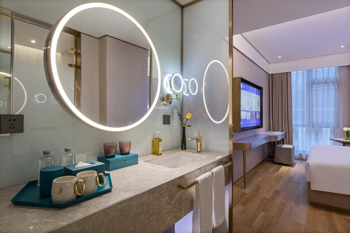 Ein modernes Hotelbad mit einem großen Spiegel, einem Schminktisch mit Fernseher und einem Bett im angrenzenden Zimmer.