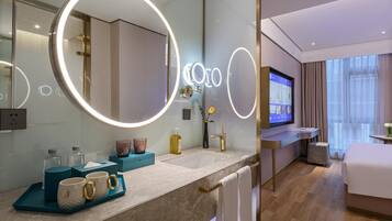 A modern hotel bathroom with a large mirror, a vanity with a TV, and a bed in the adjacent room.