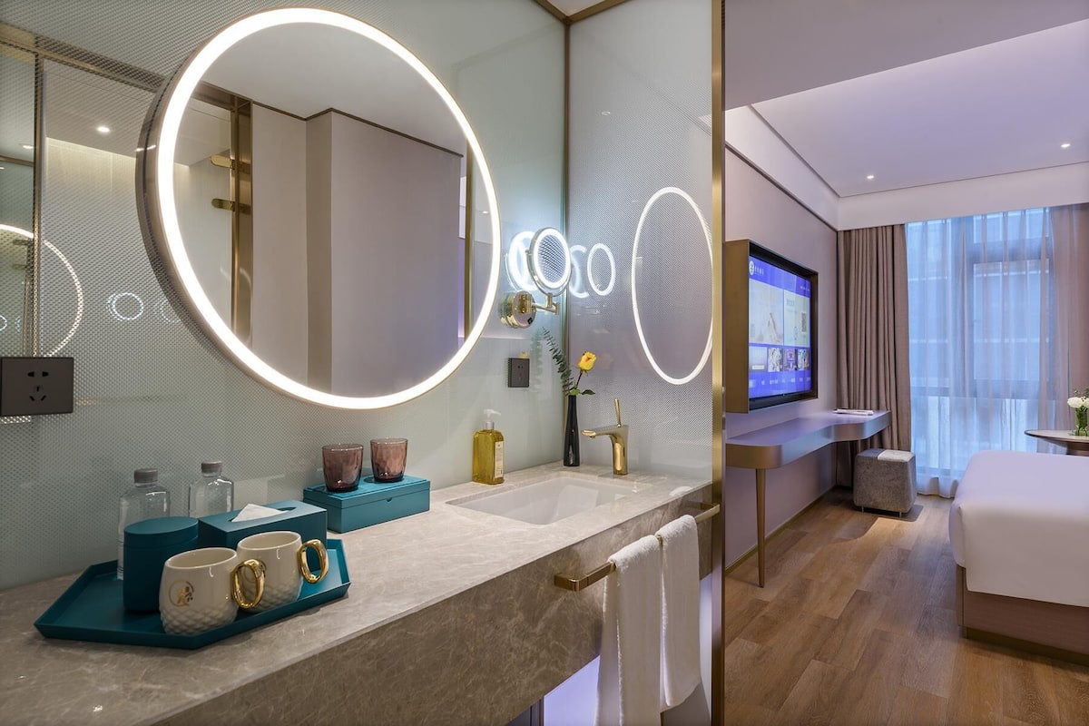 A modern hotel bathroom with a large mirror, a vanity with a TV, and a bed in the adjacent room.