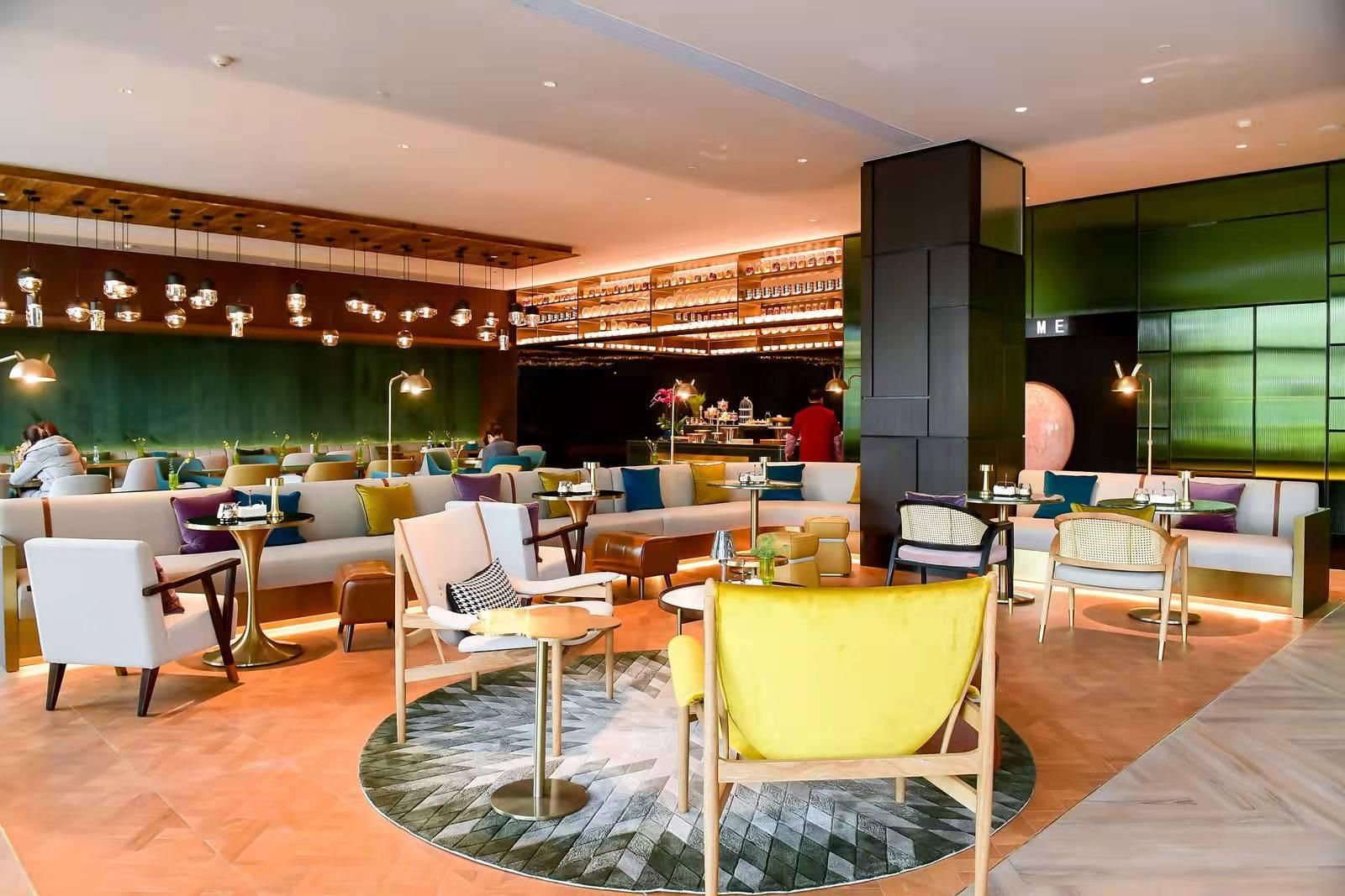 A modern lounge area with sofas, chairs, and tables, featuring a bar and a large green wall.