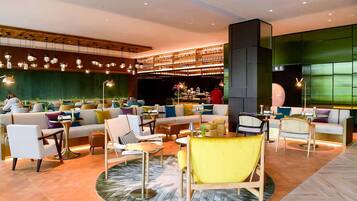 A modern lounge area with sofas, chairs, and tables, featuring a bar and a large green wall.