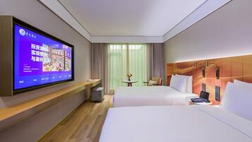 A hotel room with a large flat-screen TV mounted on the wall, a bed with white linens, a small round table with a vase of flowers, and a chair.