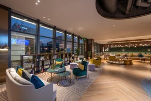 Ein moderner Loungebereich mit Sitzgelegenheiten, Tischen und großen Fenstern mit Blick auf die Stadtlandschaft.