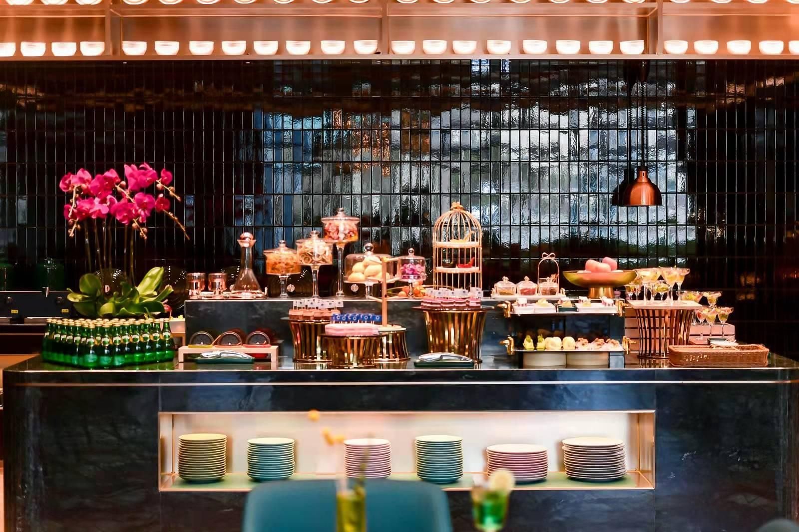 A bar area with a variety of drinks and desserts, a flower arrangement, and a view through the window.