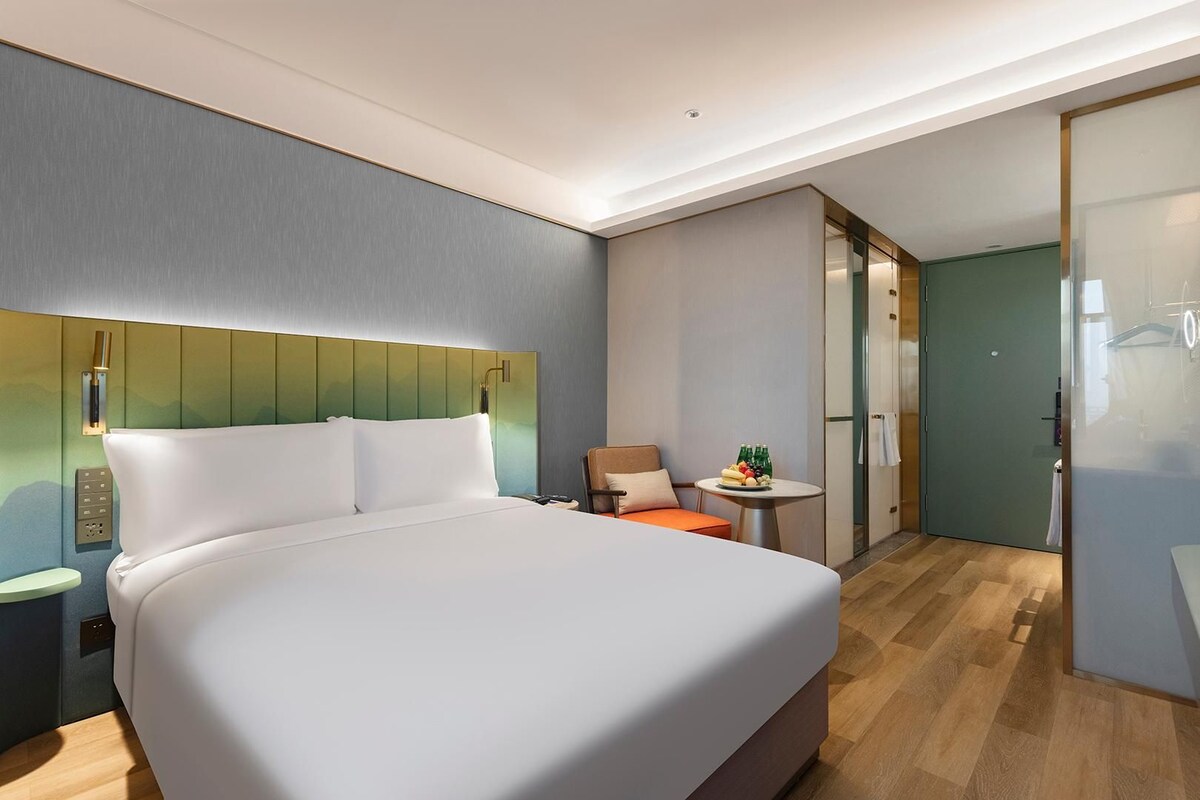 Une chambre d’hôtel moderne avec un grand lit, une petite table sur laquelle se trouve un panier de fruits, une chaise et un dressing.