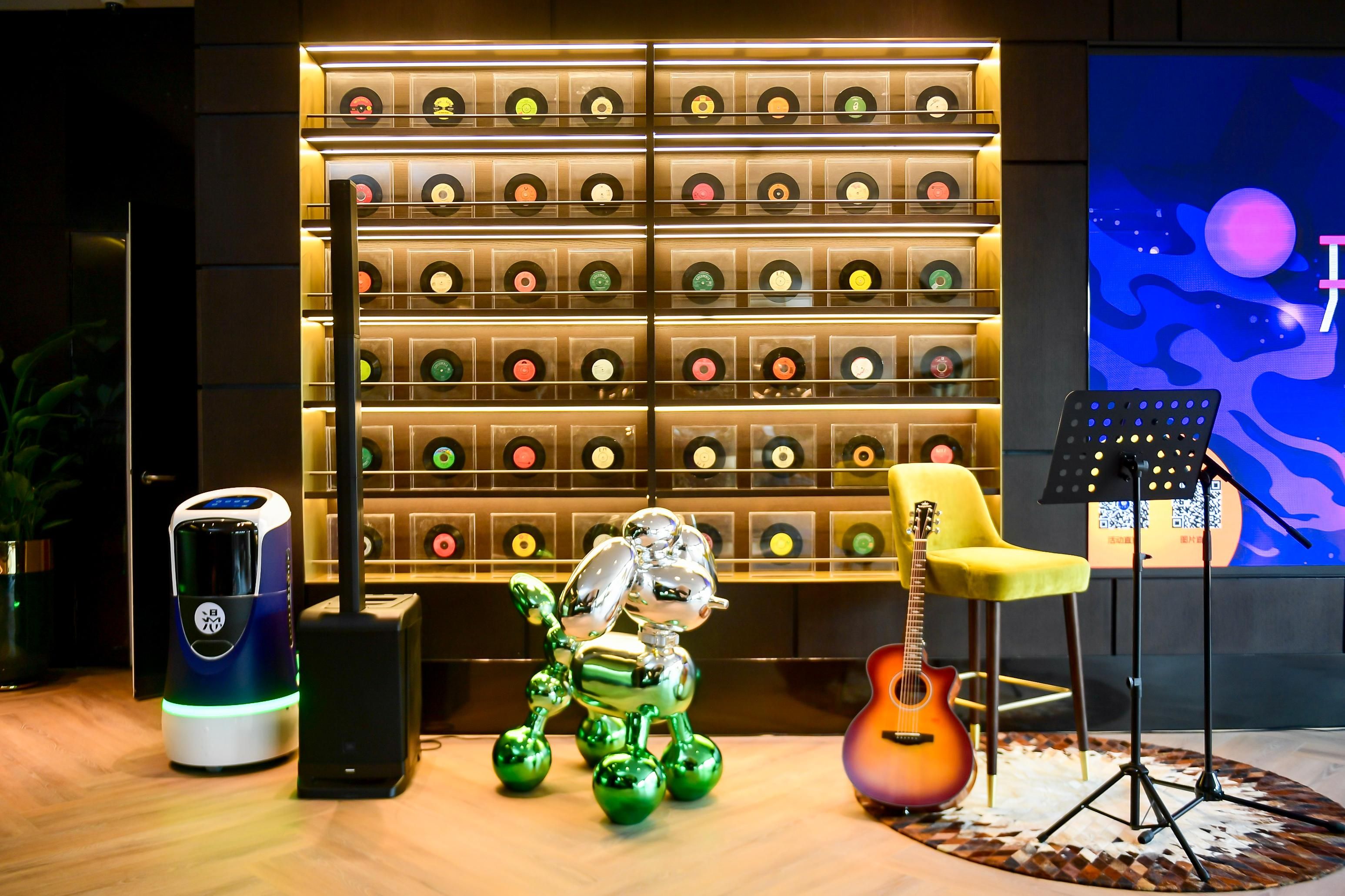 A room with a wall of vinyl records, a robot, a guitar, a speaker, and a chair.
