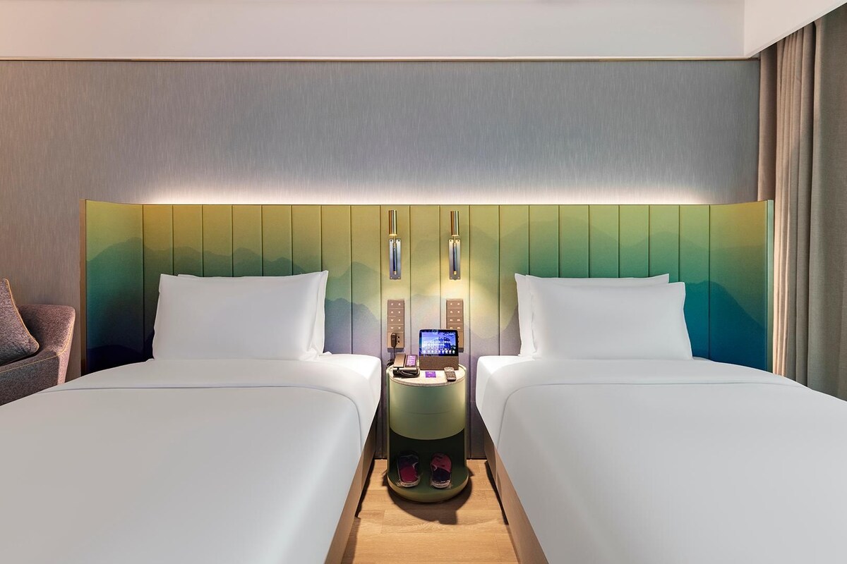 Une chambre d’hôtel avec deux lits, une table de chevet avec un téléphone et un appareil numérique, et un mur orné d’un motif à dégradé de couleurs.