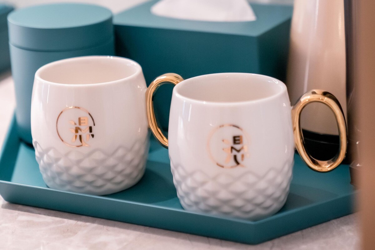 Deux tasses en céramique blanche, dotées d’anses dorées et d’un anneau doré, posées sur un plateau de couleur turquoise.