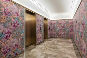 Un couloir avec deux ascenseurs, dont l’un est équipé d’un panneau de commande. Les murs sont décorés d’un motif floral.