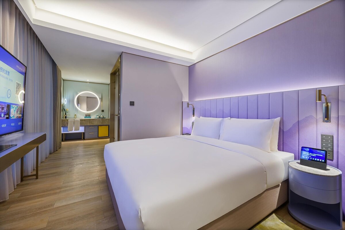 Une chambre d’hôtel moderne avec un grand lit, une table de chevet dotée d’une horloge numérique, et donnant sur la salle de bain et le couloir.