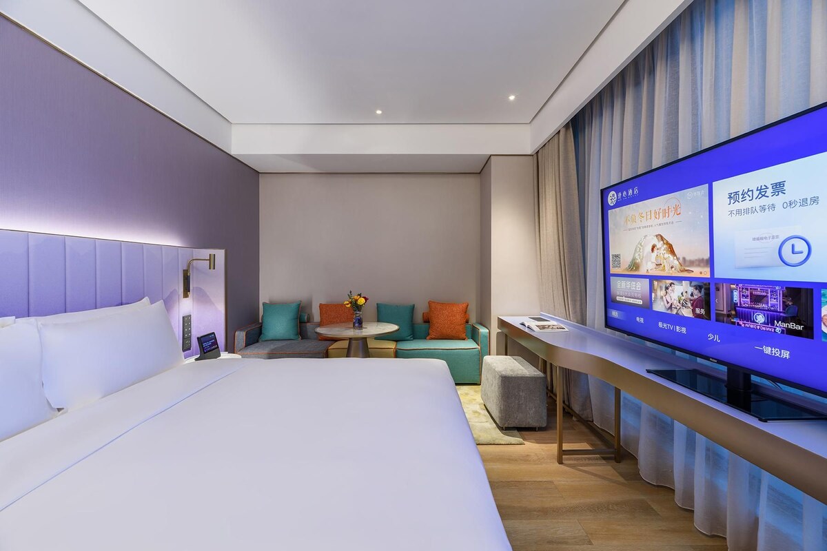 A modern hotel room with a large bed, a seating area with a small table and chairs, and a television displaying a menu.
