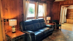 A wooden interior with a black leather sofa, two small tables with lamps, a green chair, and a view of the outdoors through a window with curtains. - Lakefront cottage on 540 acre Wall Lake / spectacular fishing & sunset views.  (Delton)