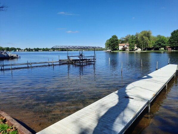 Un lago tranquilo con un muelle que se adentra en el agua, rodeado de árboles y casas al fondo.