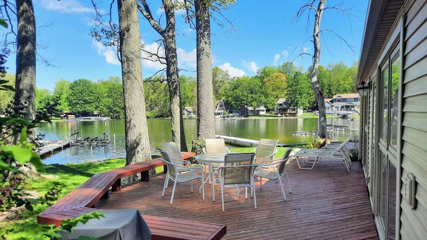 A deck with outdoor furniture overlooking a lake, surrounded by trees and houses in the background. - Lakefront cottage on 540 acre Wall Lake / spectacular fishing & sunset views.  (Delton)