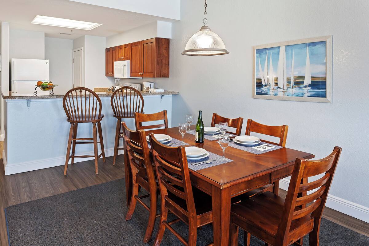 Un comedor con una mesa de madera dispuesta para seis personas, rodeada de sillas de madera. Se aprecia una cocina al fondo con armarios de madera y un refrigerador blanco.