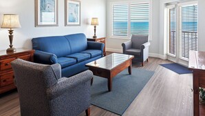 Sala de estar con un sofá azul, un sillón a juego, una mesa de centro de madera y una alfombra azul. Hay dos lámparas sobre mesitas de noche, y la habitación tiene vistas a través de las persianas.