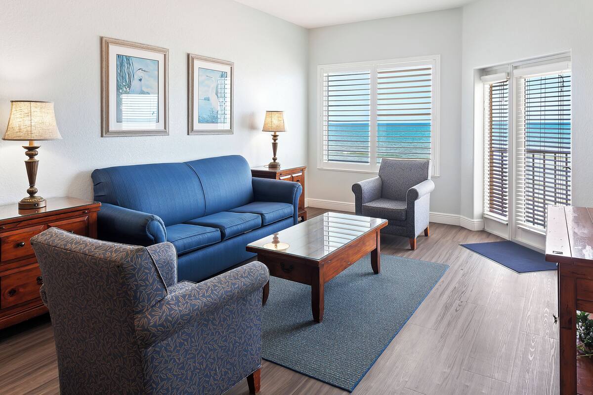 Sala de estar con un sofá azul, un sillón a juego, una mesa de centro de madera y una alfombra azul. Hay dos lámparas sobre mesitas de noche, y la habitación tiene vistas a través de las persianas.