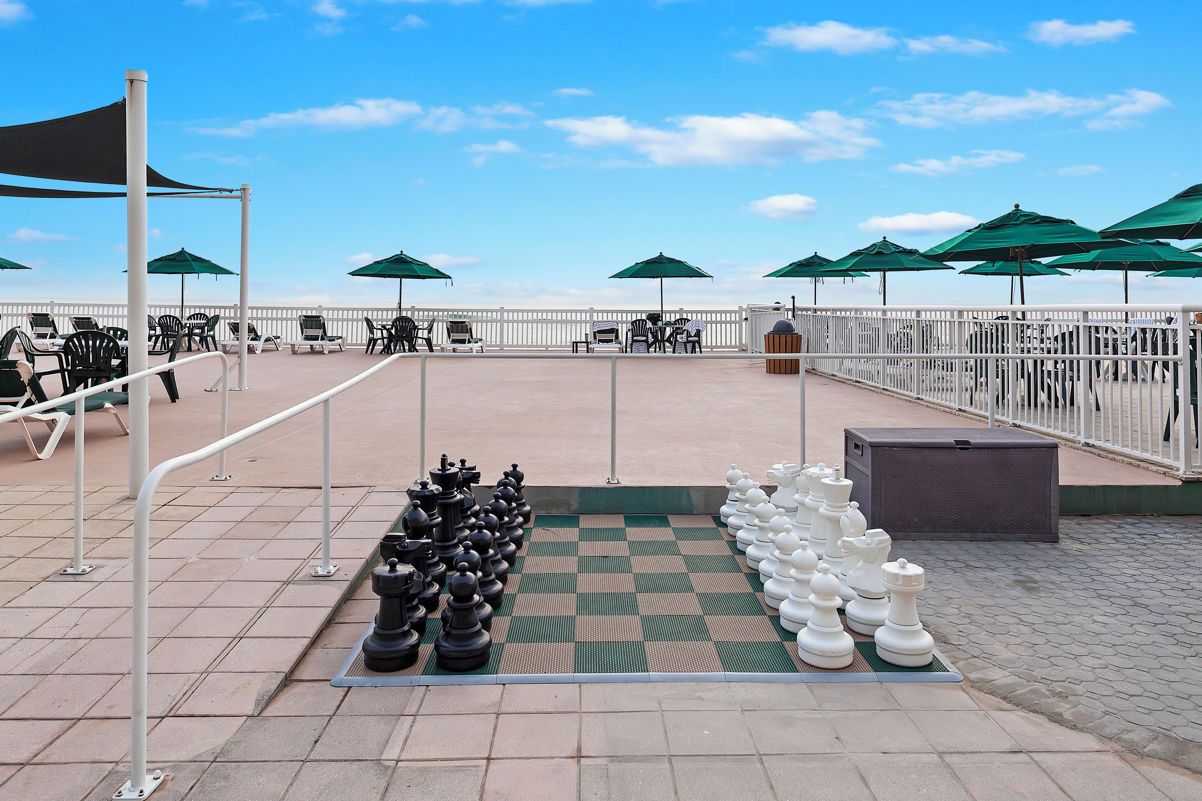 A large outdoor chessboard set up on a terrace with tables and chairs, surrounded by umbrellas and railings.