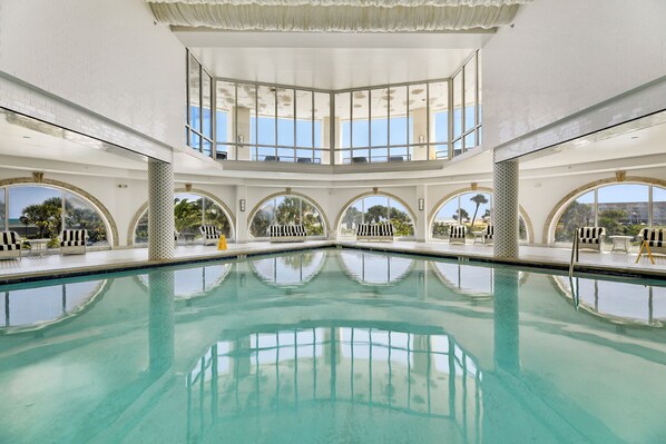 A large indoor swimming pool with multiple levels of seating and large arched windows.