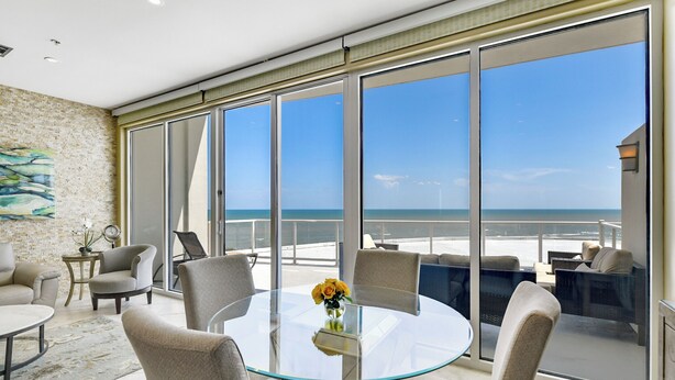 Een moderne kamer met een glazen tafel, beige stoelen en uitzicht op de oceaan door grote ramen.