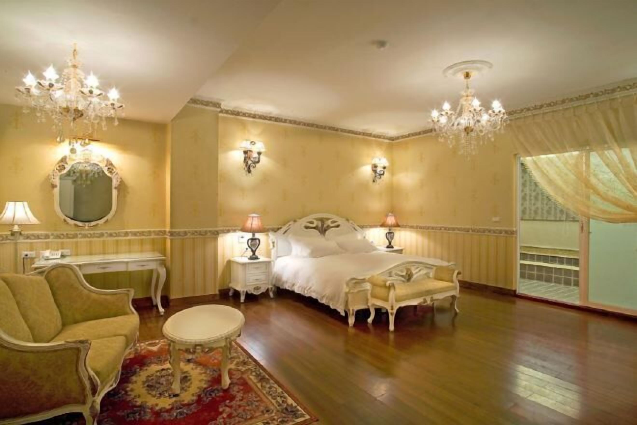 A spacious bedroom with a large bed, two nightstands, a chandelier, a mirror, and a chaise lounge.