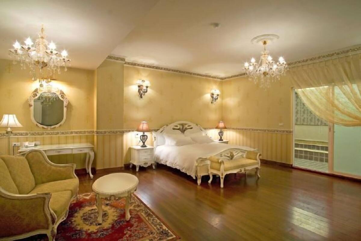 A spacious bedroom with a large bed, two nightstands, a chandelier, a mirror, and a chaise lounge.