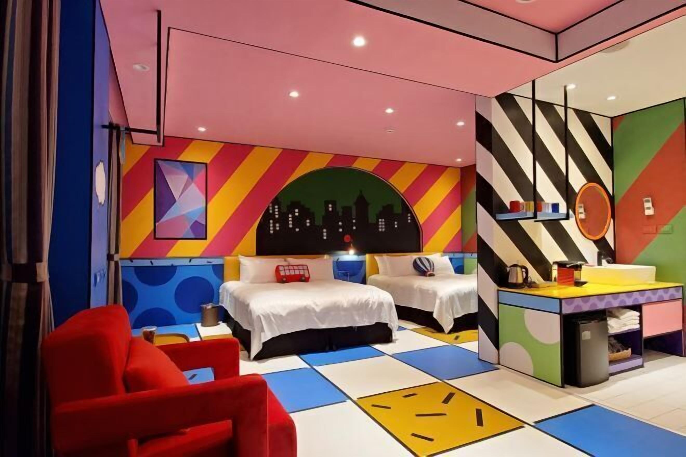 A colorful hotel room with two beds, a red chair, a cityscape mural, and a mirror with a striped pattern.