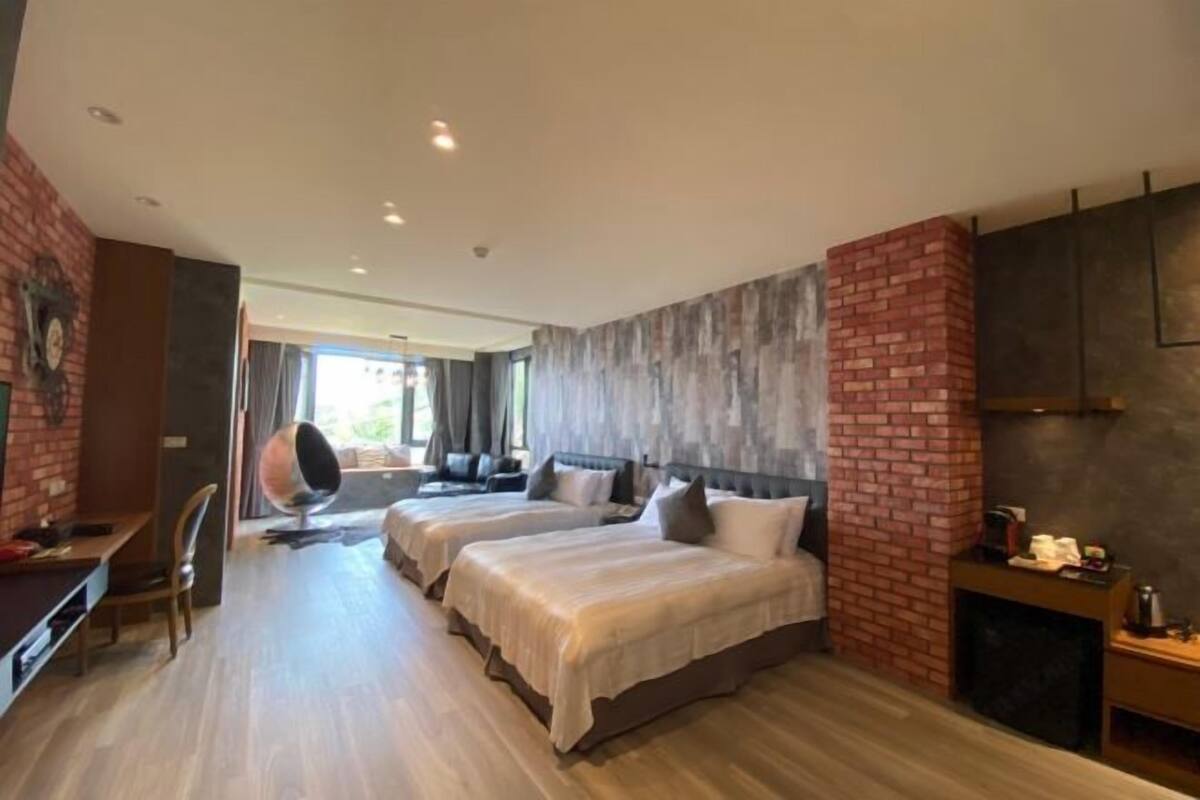 A hotel room with two beds, a brick accent wall, a seating area with a chair and a round table, and a view of the outdoors.