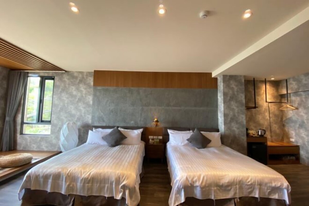 Quarto de hotel com duas camas, cabeceira de madeira, mesa de cabeceira, luminária e janela com cortinas.
