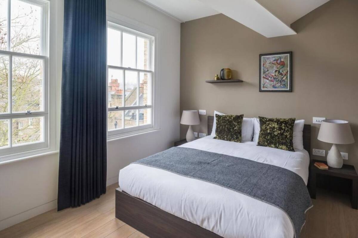 A bedroom with a large bed, two bedside tables with lamps, a window with curtains, a painting on the wall, and a view of buildings outside.