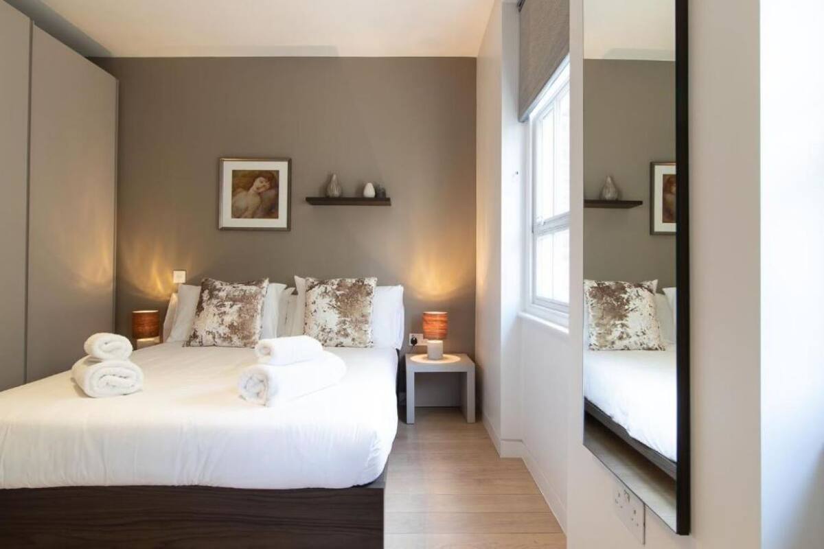 A hotel room with a large bed, a bedside table with a lamp, a small shelf with decorative items, and a large mirror on the wall.