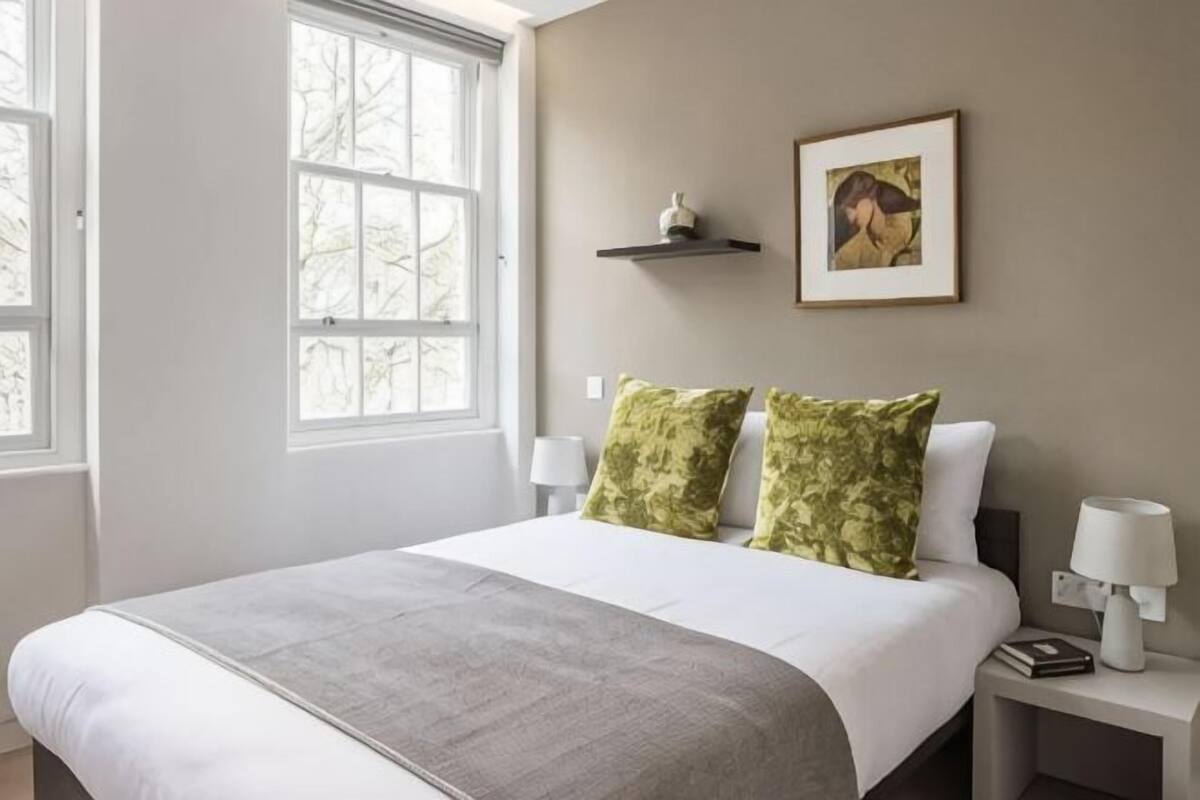 A bedroom with a bed, two pillows, a bedside table with a lamp, a nightstand with a lamp, a framed picture on the wall, and a window.