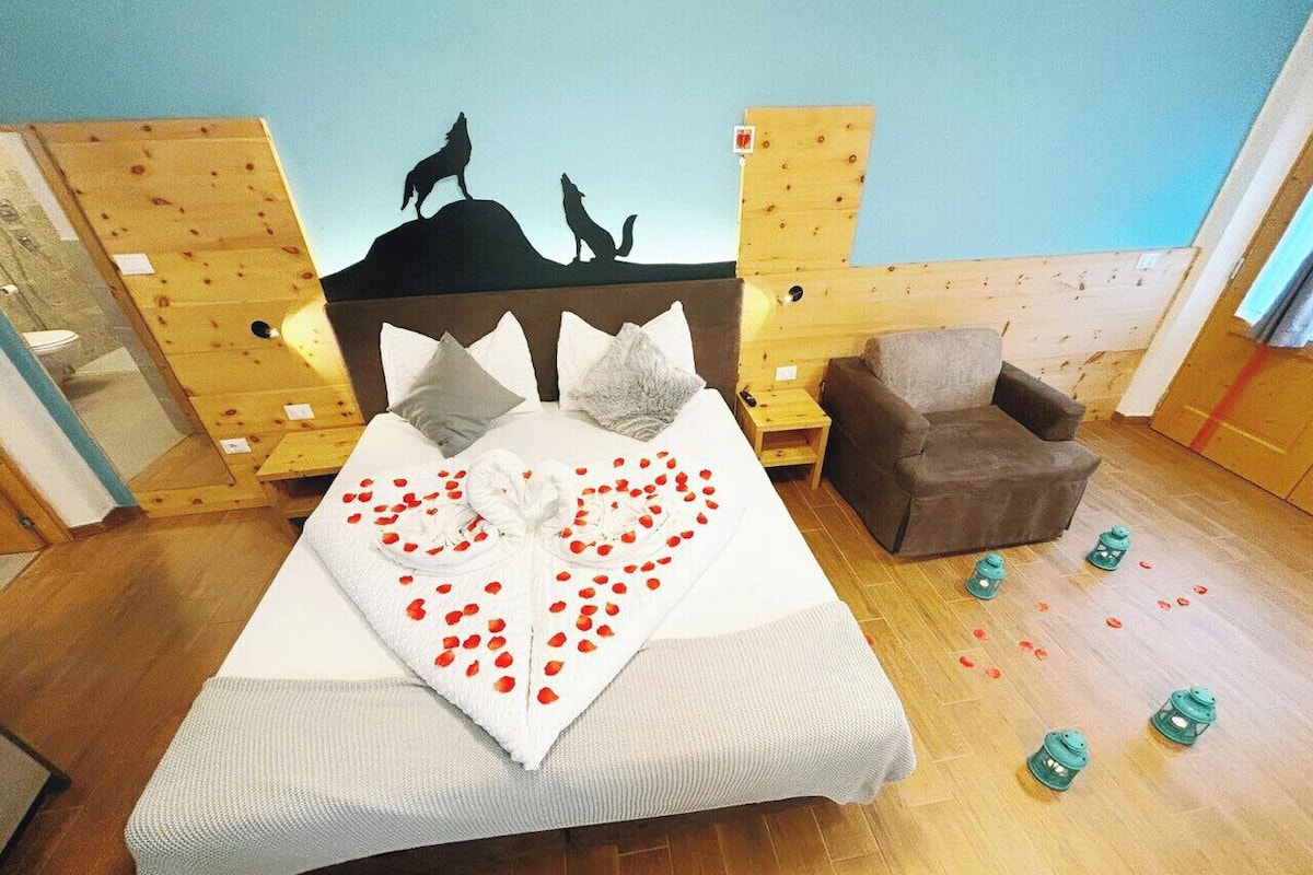 A bedroom with a bed, a heart-shaped arrangement of rose petals, a wooden headboard, a brown armchair, and a wall mural of wolves.
