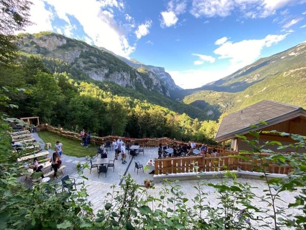 Een terras buiten met tafels en stoelen, omringd door weelderige begroeiing en bergen op de achtergrond.