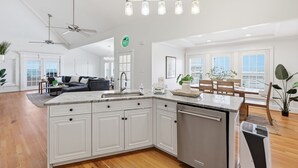 A spacious kitchen with white cabinetry, a central island, and a dining area with wooden chairs and a table. - Beachfront, Large Deck, Sleeps 9, Open, Airy & Spacious (Saint Helena Island)