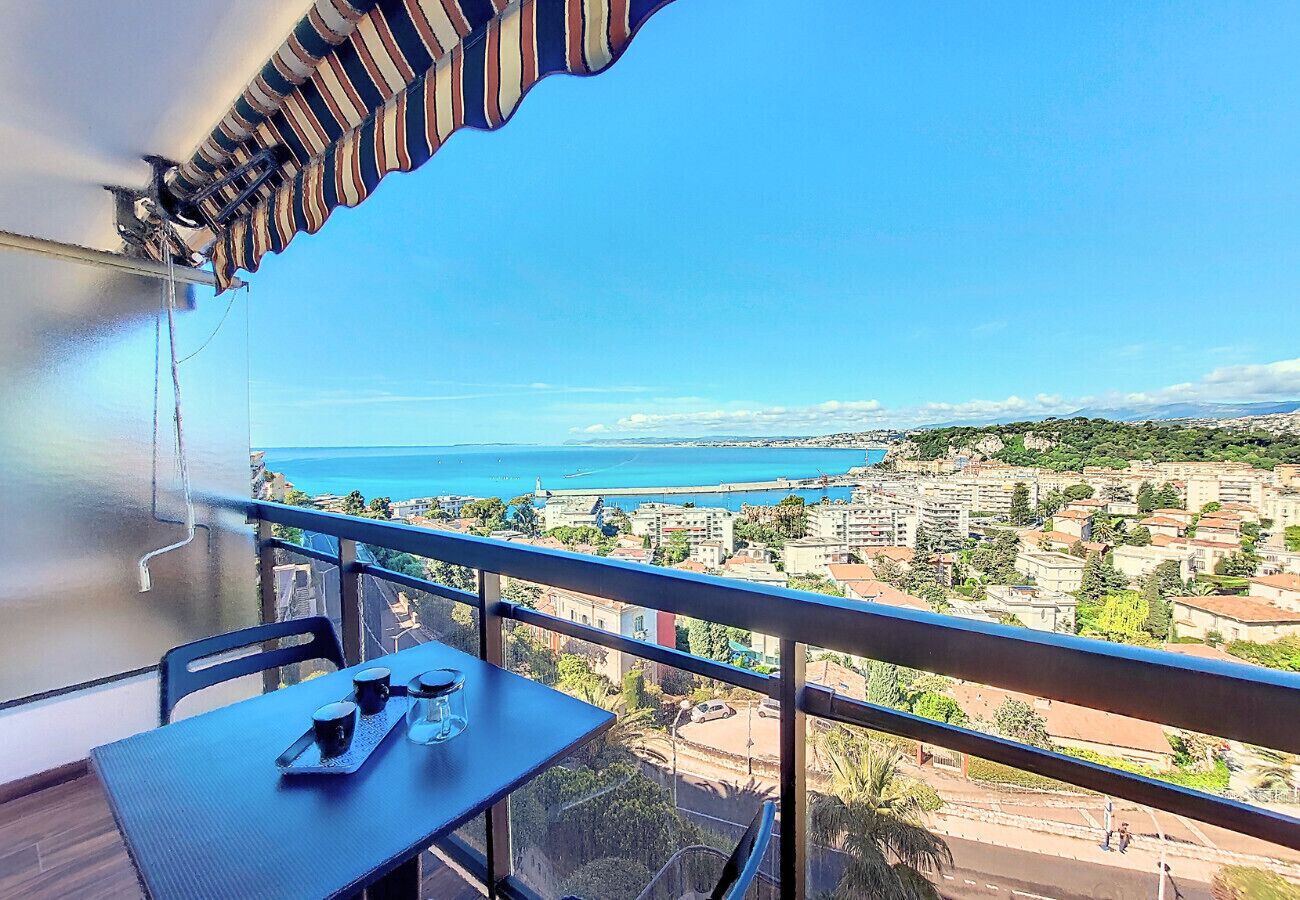 A balcony with a table and chairs, overlooking a coastal cityscape with buildings and a view of the sea.