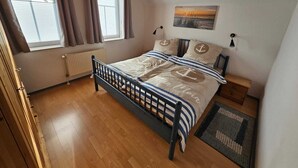 A bedroom with a wooden floor, a bed with striped bedding, a painting on the wall, and a window with curtains. - Ferienhaus Meeresbrise in Nessmersiel - NEU in der Vermietung (Dornum)