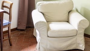 Un sillón blanco con una silla de madera al lado, una cortina marrón y un suelo de baldosas.