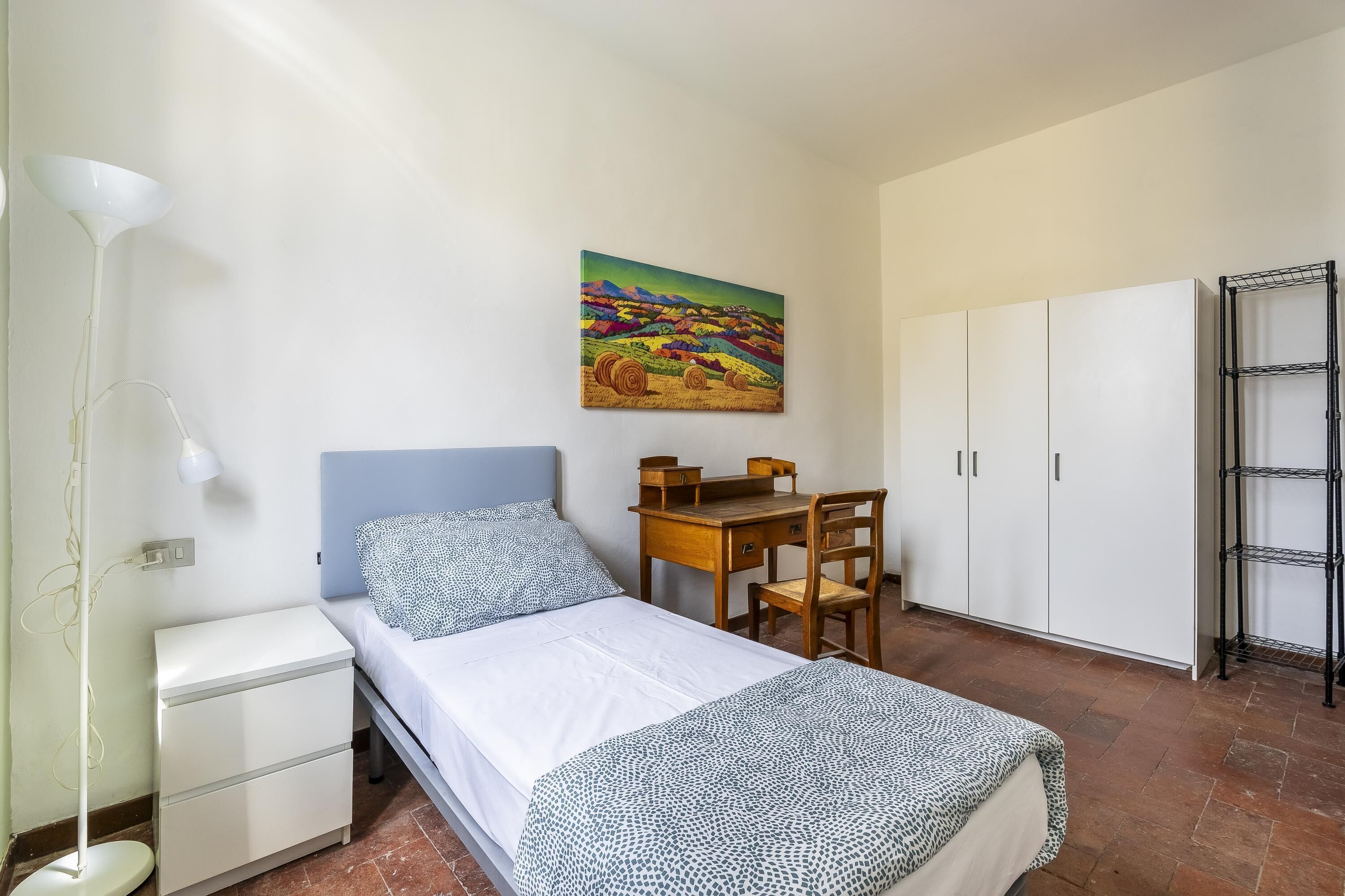 Un dormitorio con cama, escritorio, silla, armario y un cuadro en la pared.