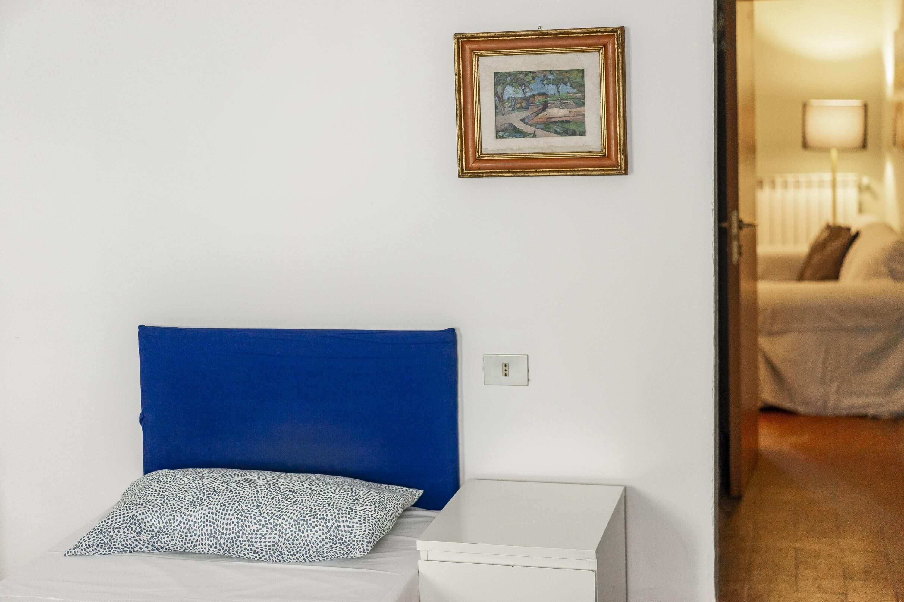 A room with a bed, a bedside table, a framed picture on the wall, and a view into another room.