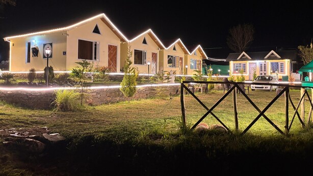 Una hilera de casas de campo amarillas con detalles blancos y luces de cadena, rodeadas de césped y un muro de piedra.