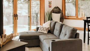 Un salon moderne avec un canapé d’angle, une table basse et un coin repas avec une chaise. De grandes fenêtres laissent entrer la lumière naturelle et offrent une vue sur l’extérieur.